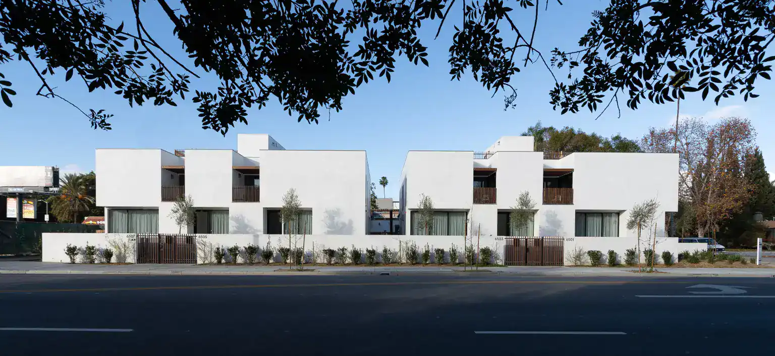 Contemporary white residential buildings with minimalist cubic forms. Wooden balcony railings and large glass doors enhance modern clarity. Sleek landscaping with small trees lines the perimeter.
