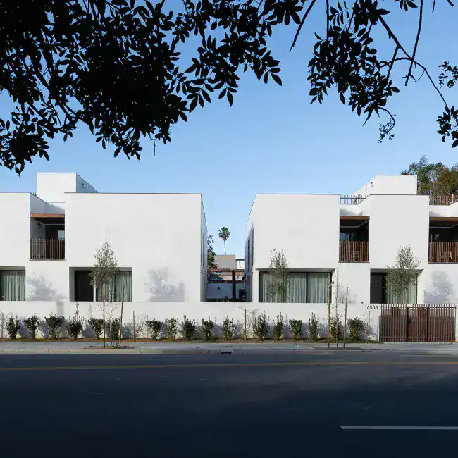 Contemporary white residential buildings with minimalist cubic forms. Wooden balcony railings and large glass doors enhance modern clarity. Sleek landscaping with small trees lines the perimeter.