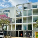 Modern white multi-story apartment building with open-air corridors and balconies, featuring vertical slat accents and a geometric facade.