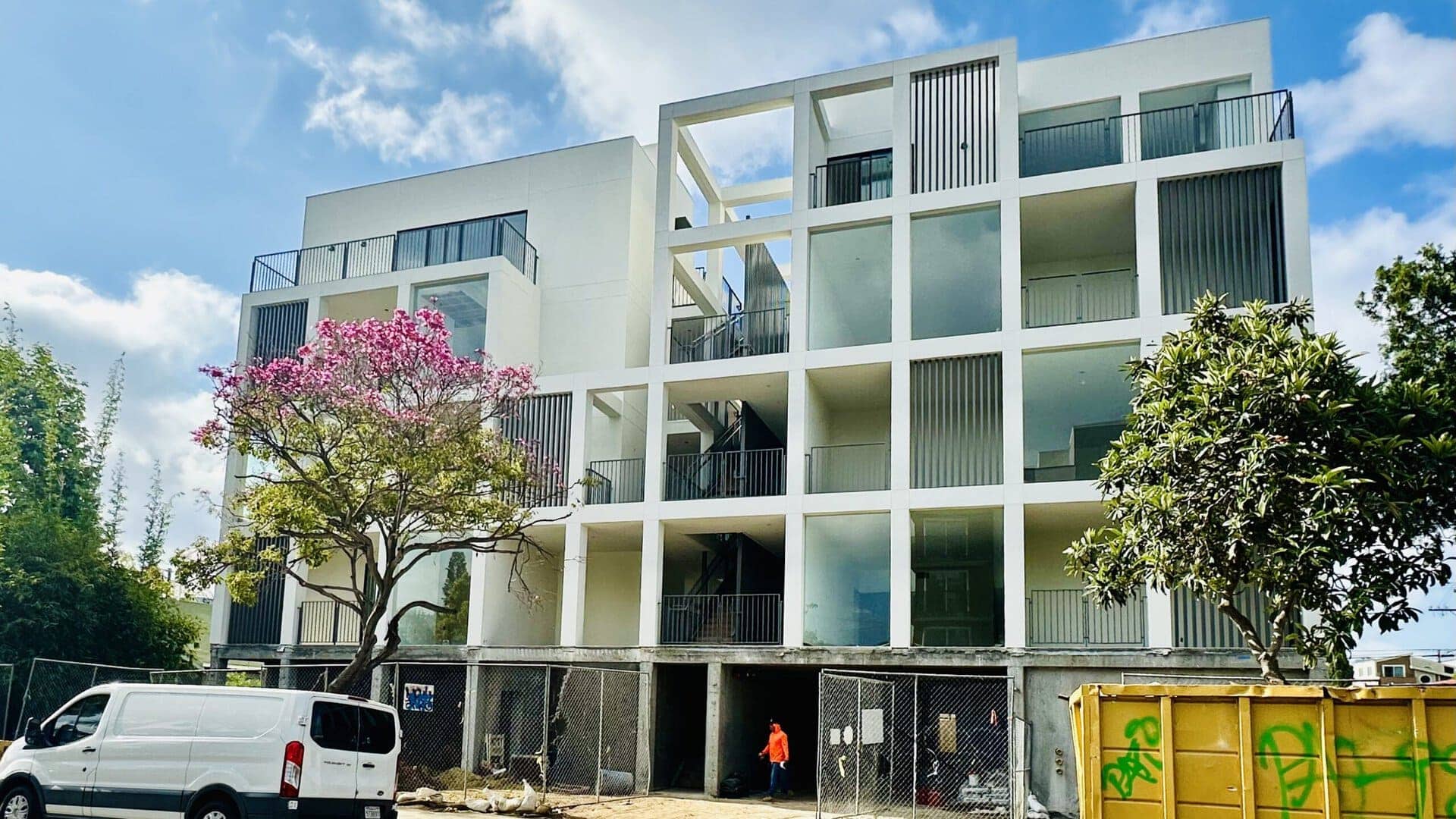 Modern white multi-story apartment building with open-air corridors and balconies, featuring vertical slat accents and a geometric facade.