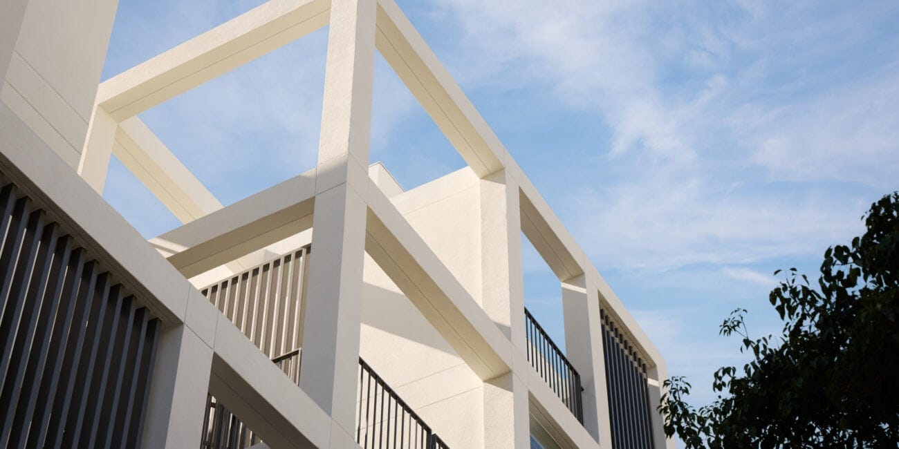 Modern white concrete building with large geometric open frames and black metal balcony railings under a blue sky.