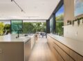 Contemporary kitchen with minimalist design, white surfaces, natural wood cabinetry, expansive glass sliding doors showing lush greenery outside.