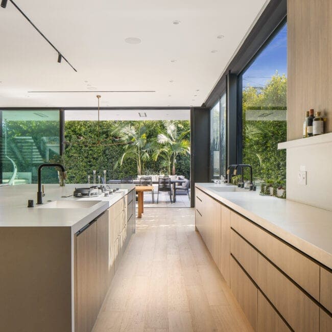 Contemporary kitchen with minimalist design, white surfaces, natural wood cabinetry, expansive glass sliding doors showing lush greenery outside.