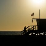 Silhouette of a lifeguard tower with an American flag on a beach during sunset.