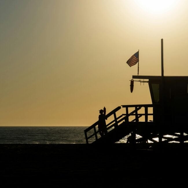 Silhouette of a lifeguard tower with an American flag on a beach during sunset.