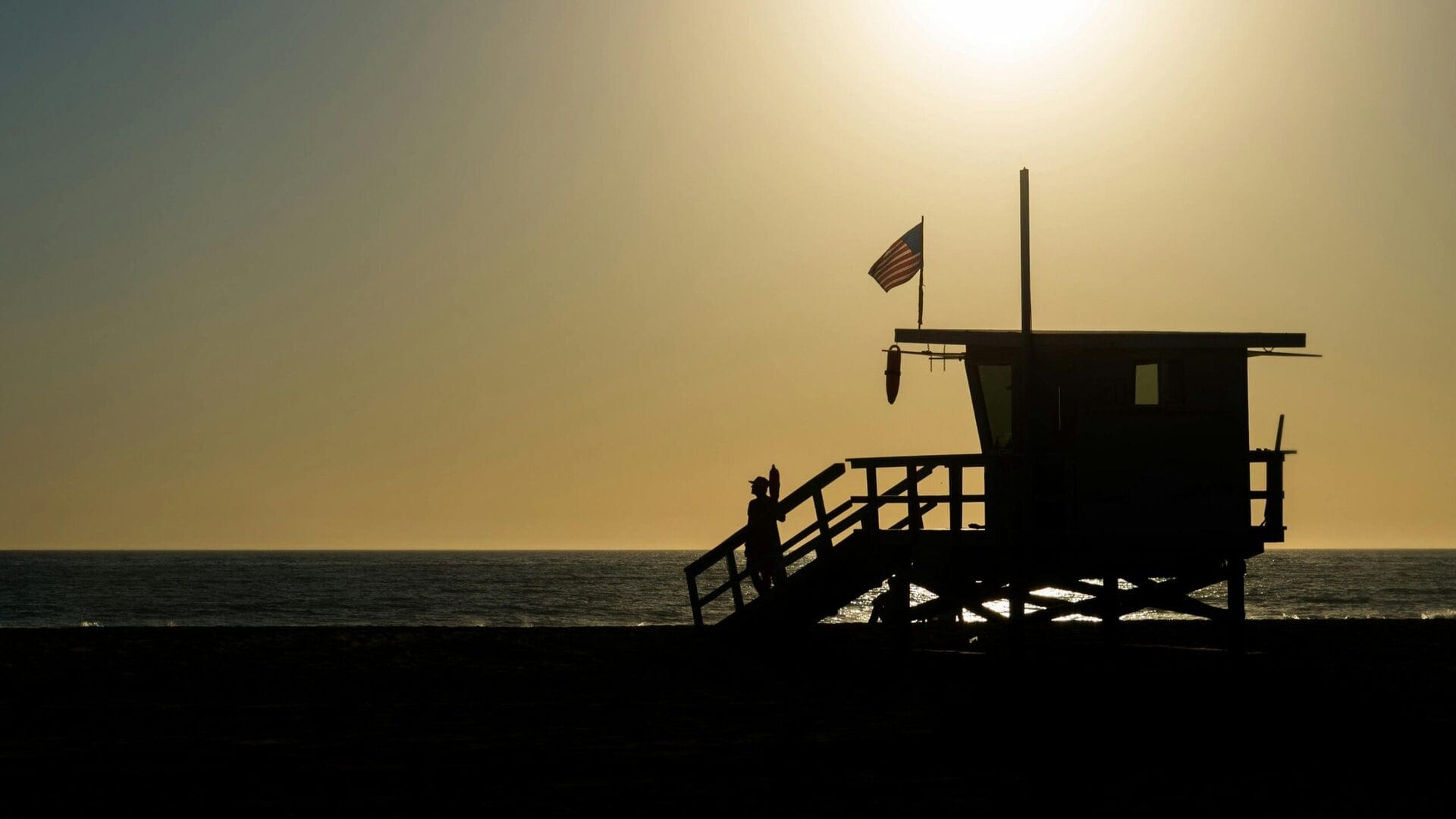Silhouette of a lifeguard tower with an American flag on a beach during sunset.